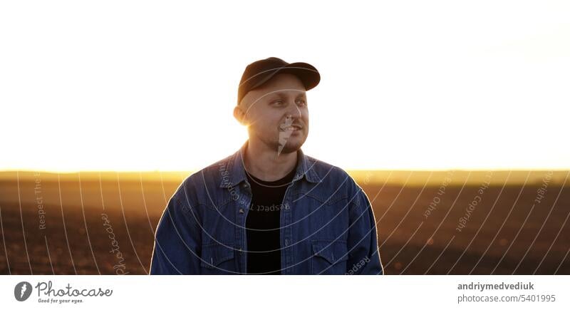 Portrait of smiling male farmer in cap is walking through endless plowed farmland field on sunset. Professional agronomist inspecting cultivated land in sunlight. Agribusiness concept and farming.