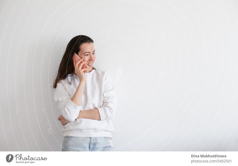 Young woman holding a mobile phone, smiling, using social media. Millennial person in white sweatshirt on white background. People using technology. Work call.