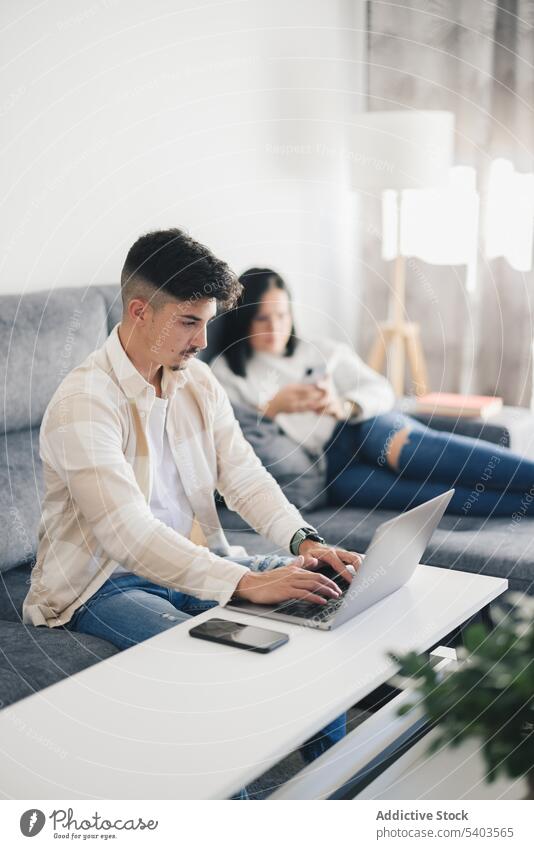 Young man working on sofa with laptop near woman with mobile phone in living room apartment brunette caucasian computer couple entertainment female furniture