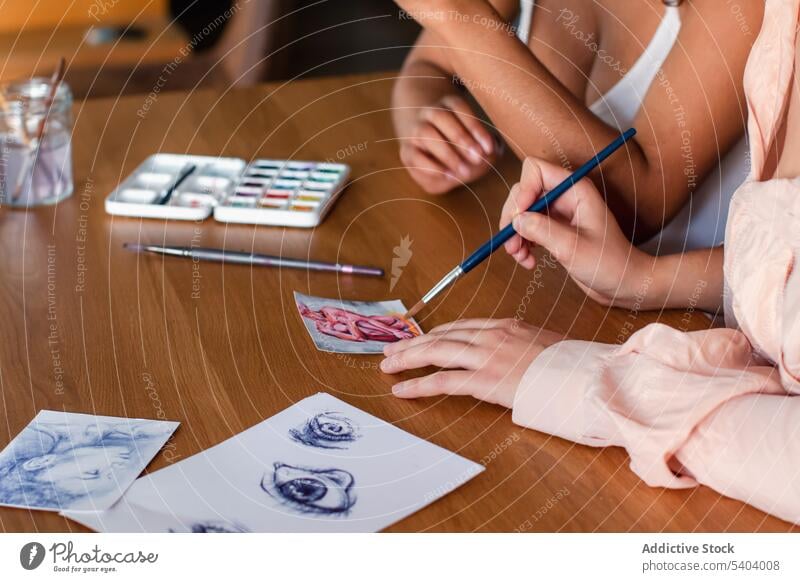 Side view of crop anonymous diverse lesbian couple drawing with paintbrushes on paper on wooden table hobby together homosexual artist relationship multiethnic