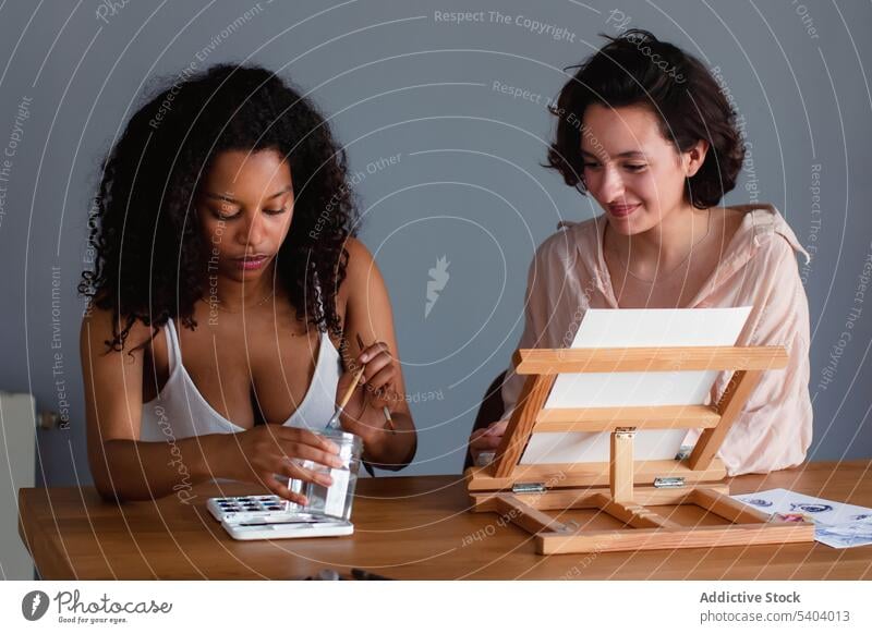 Focused diverse women sitting at table with painting brush and water colors friend mix concentrate smile watercolor young female african american black