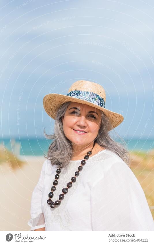 Smiling elderly lady on sunny beach woman summer seashore coast portrait content white dress travel smile vacation seaside aged style ocean straw hat female