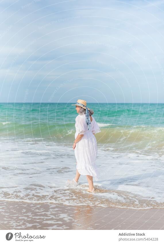 Woman walking on sandy beach woman freedom enjoy vacation summer sea nature elderly senior pensioner aged shore wave water seashore coast female ocean straw hat