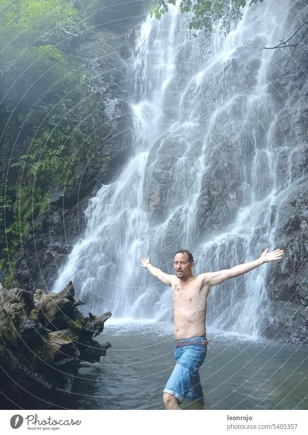 A man stands in front of a waterfall with his upper body exposed and his arms outstretched. He stands knee-deep in the water and next to him is an old tree trunk covered with moss. He is happy and grateful to be in nature!