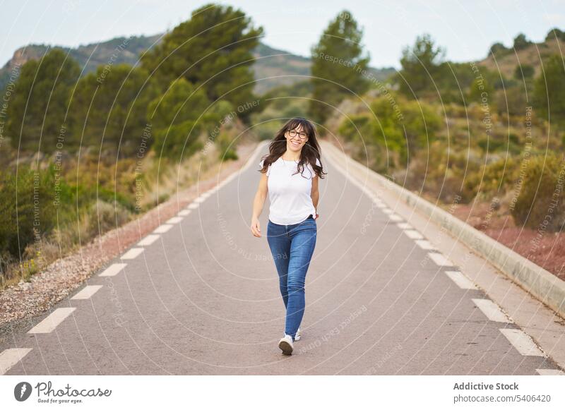 Happy woman walking along road tourist traveler stroll pedestrian asphalt way female countryside hispanic trip tourism smile lady cheerful wanderlust path