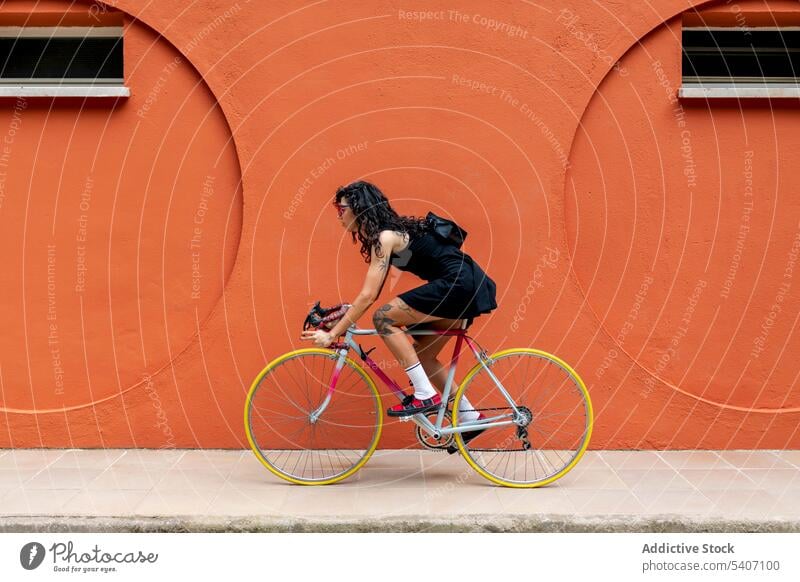 Young ethnic woman with handbag riding bicycle on sidewalk near orange wall street ride building sneakers yellow tire female young style sunglasses urban city