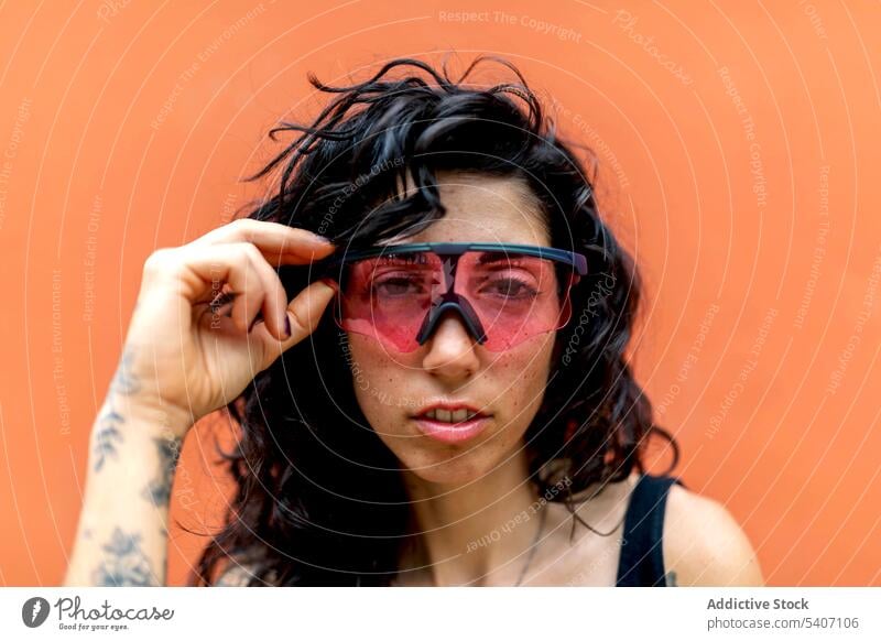 Stylish young ethnic woman touching sunglasses with hand in daylight portrait smile happy orange wall tattoo summer female hispanic daytime feminine style