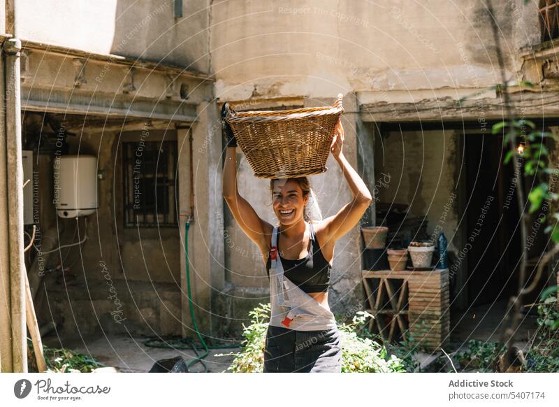 Happy young woman carrying wicker basket on head near green plants in house garden smile building shabby positive summer female enjoy glad happy cheerful