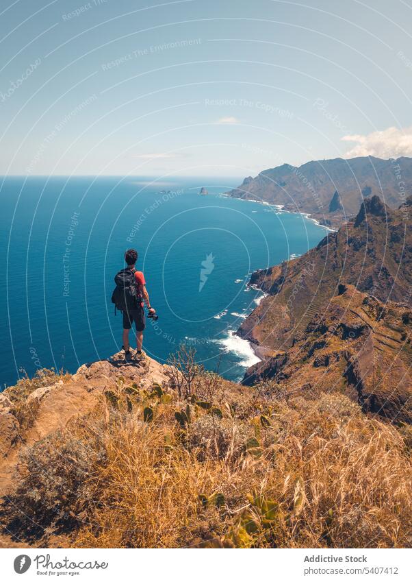 Young man with backpack standing on cliff with camera admiring seascape mountain traveler hiker nature landscape adventure male trip tourism vacation tenerife