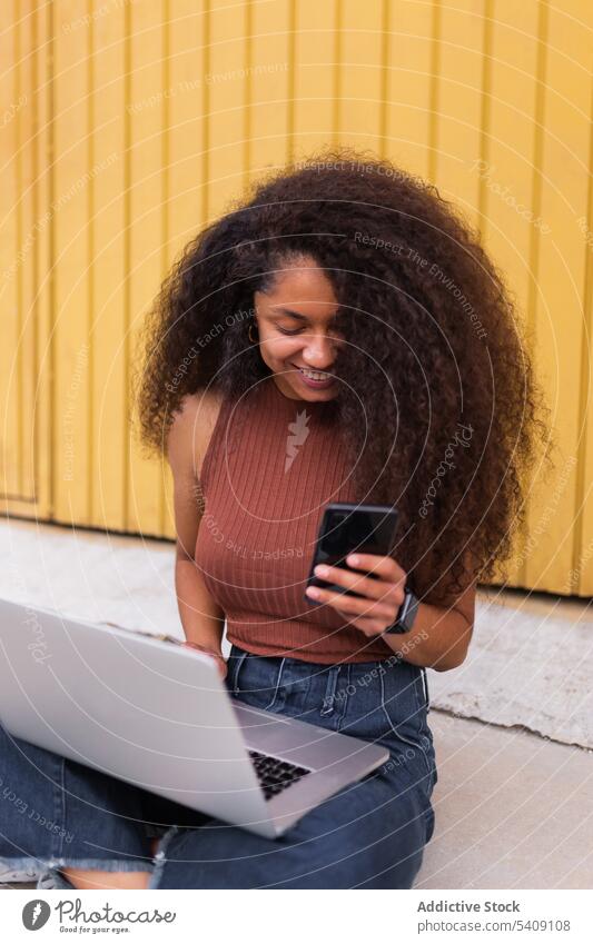 Cheerful African American self employed woman sitting on street with laptop smartphone freelance curly hair city entrepreneur ethnic black african american