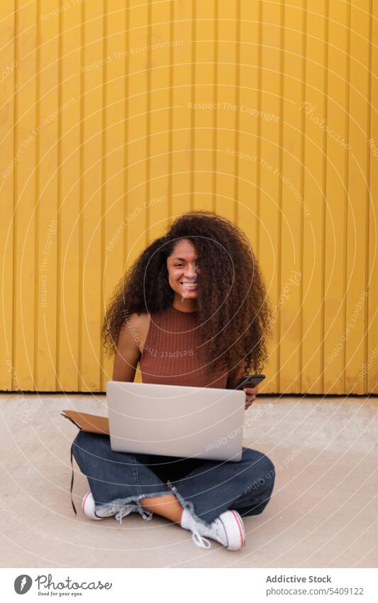 Cheerful African American self employed woman sitting on street with laptop smartphone freelance curly hair city entrepreneur ethnic black african american