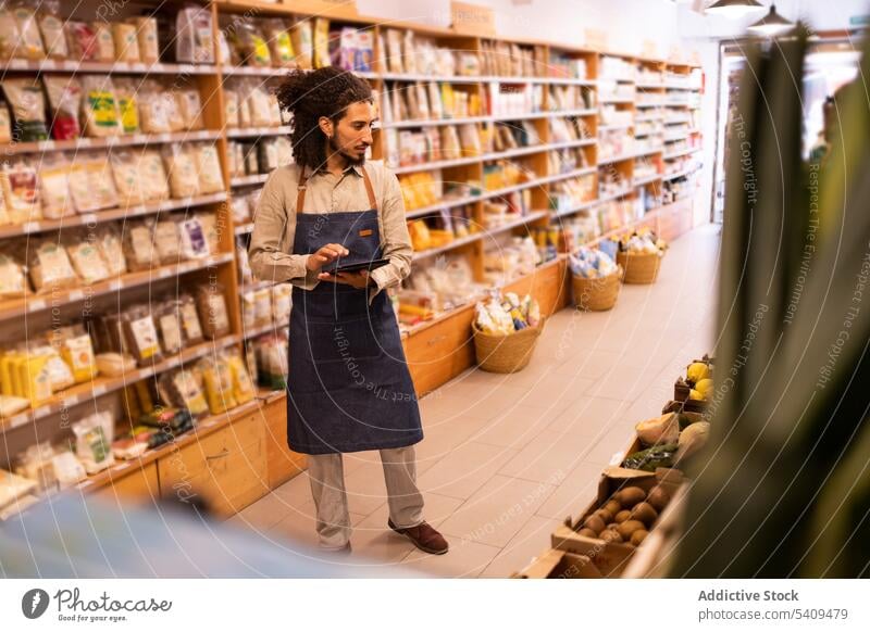 Serious man with tablet checking goods in grocery store shop supermarket inventory control serious food gadget curly hair focus shelf using device stand modern