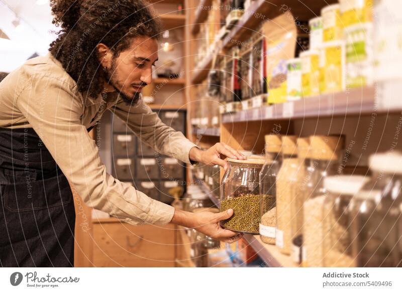 Ethnic male worker with jar of bulk standing in supermarket man seller food grocery store job service shelf young ethnic curly hair apron glass weight commerce