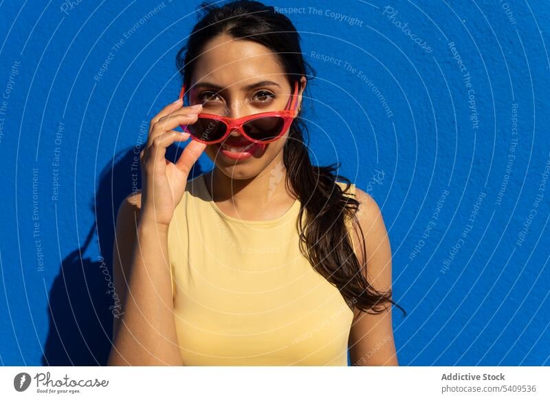 Happy young ethnic woman with sunglasses standing in light against blue wall smile shadow portrait room appearance positive happy female charming cheerful