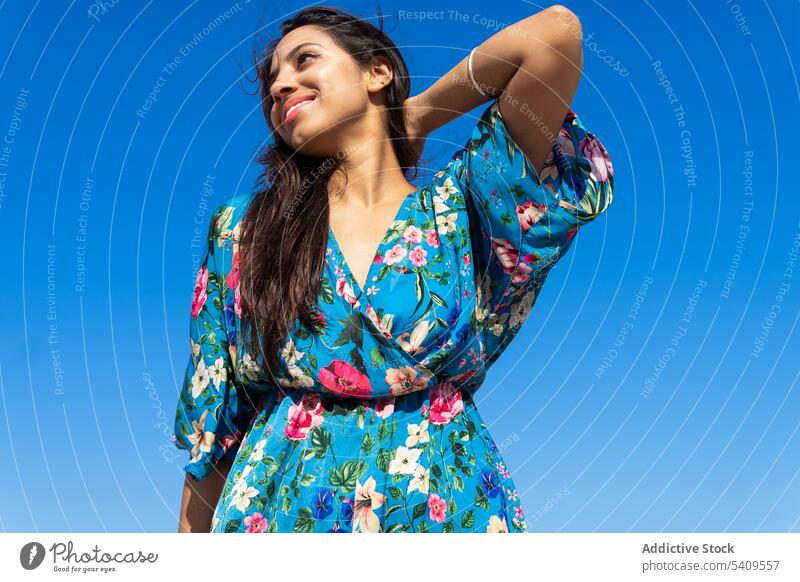 Smiling young ethnic woman standing and touching hair against blue cloudless sky smile positive blue sky happy long hair glad pleasant touch neck female