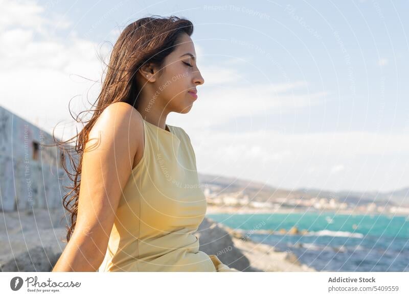 Young ethnic woman sitting on beach in daylight with eyes closed near blurred seawater smile coast nature long hair calm lean on hand shore young female indian