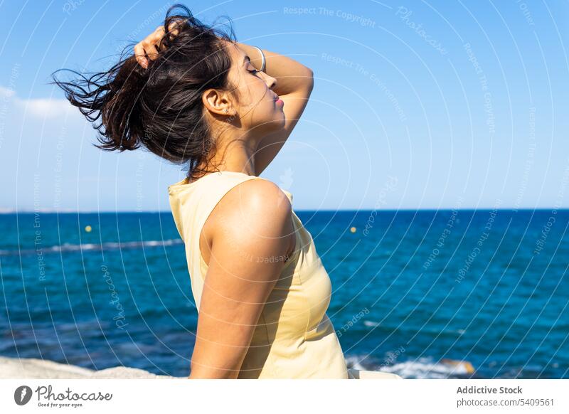 Young ethnic woman sitting on beach in daylight with eyes closed near blurred seawater coast nature long hair calm lean on hand shore young female indian rocky