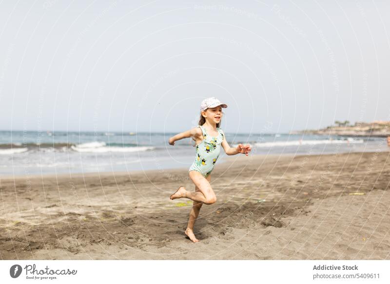 Happy kid in swimwear and cap running on sandy beach child wet barefoot smile seashore carefree summer girl preteen happy vacation wave blue sky seawater