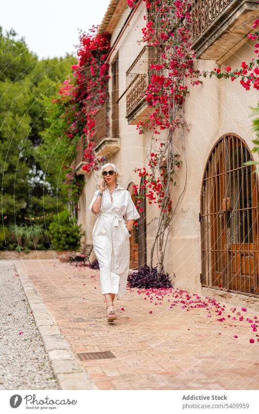 Happy senior woman with hand in pocket walking on sidewalk near building and speaking on smartphone smile plant entrance positive door female happy exterior