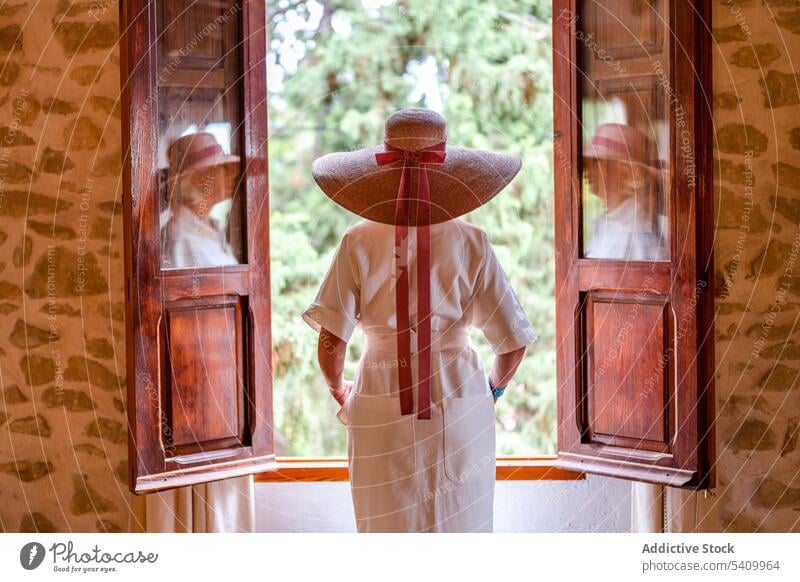 Anonymous woman in wide straw hat standing near window with glass doors reflection ribbon green tree bow tie park summer female style nature lady headwear