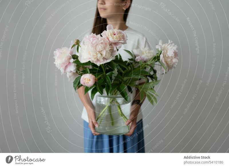 Unrecognizable preteen kid standing with blooming peony flowers in glass jar child fresh green leaf stem arrange floral blossom dress style vase daylight