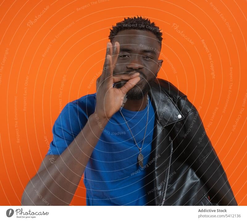 Serious young black man sitting and showing OK sign near orange wall ok gesture confident portrait serious self assured leather jacket necklace male curly hair