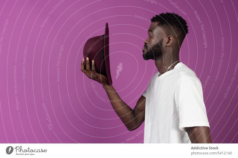 Young black man with curly hair standing with hat near purple wall confident appearance individuality serious thoughtful calm cool male young african american