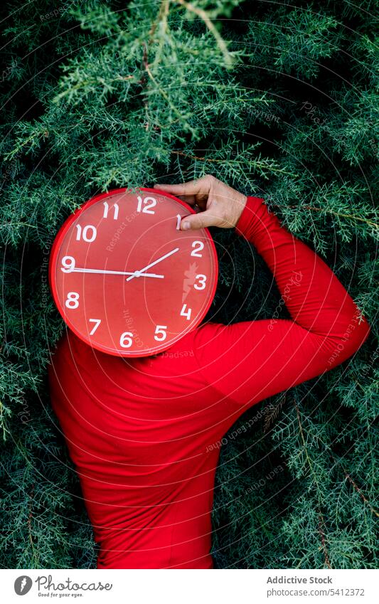 Man hiding face with red clock against green bushes man cover face hide time park nature deadline punctual male casual plant shrub garden minute daytime foliage