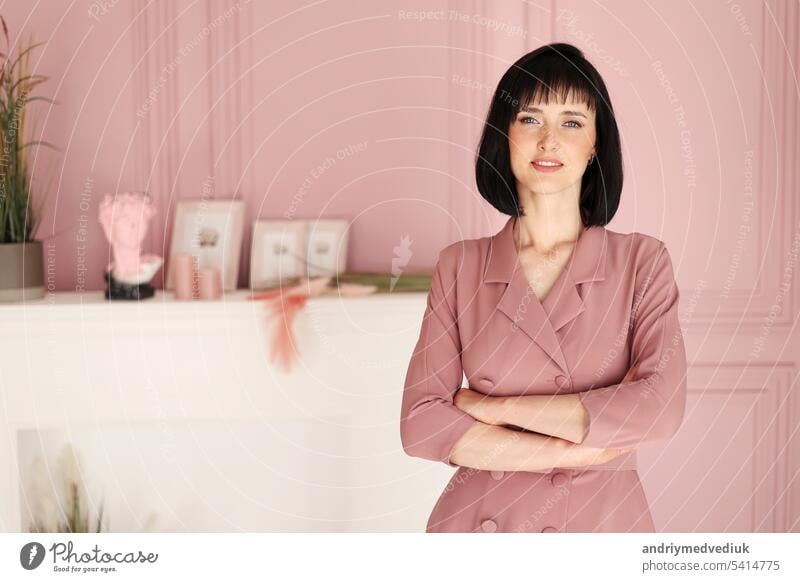 Portrait of attractive smiling brunette with short hair woman folded arms staying alone at stylish interior indoors. Female in soft pink dress is looking in camera