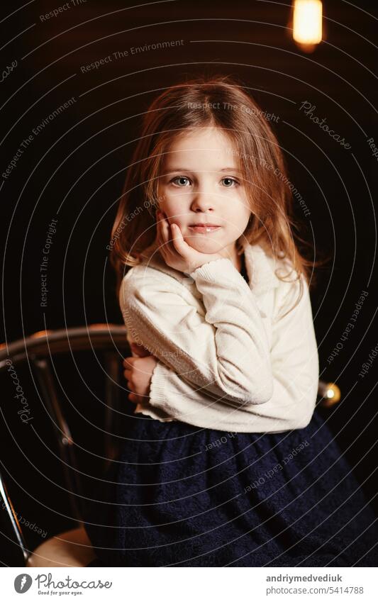 Little adorable smiling girl is sitting on metal chair with a dark background with retro light bulbs. Child in lace blue dress and knit sweater looking in camera. Happy childhood