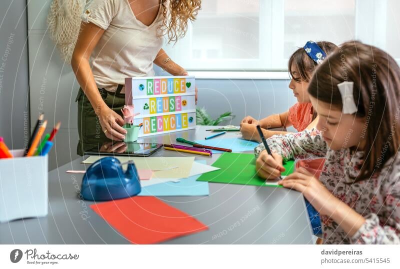 Teacher holding lightbox with text reduce, reuse, recycle while students drawing in classroom teacher unrecognizable female showing ecology cardboard crafts