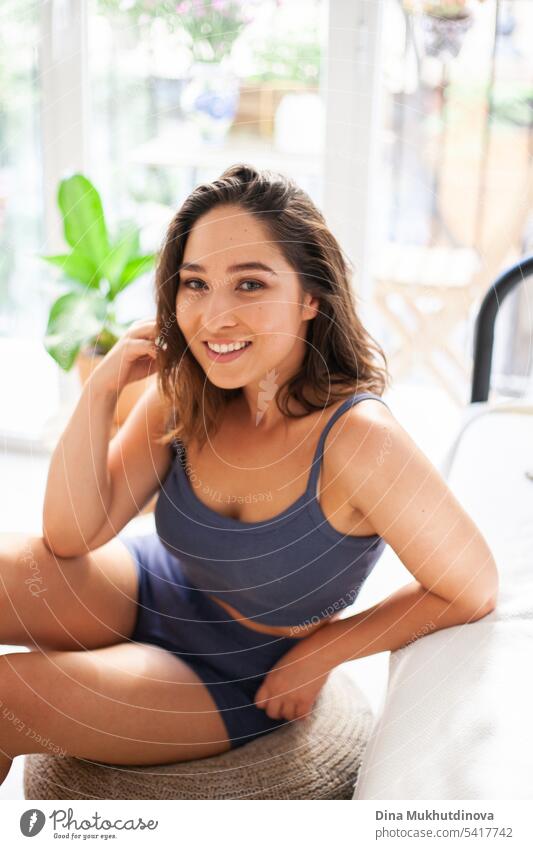 Happy young woman in minimal apartment lifestyle photo, smiling. Millennial woman at home. Interior shot with panoramic window. Girls pajama party. Good morning mood.