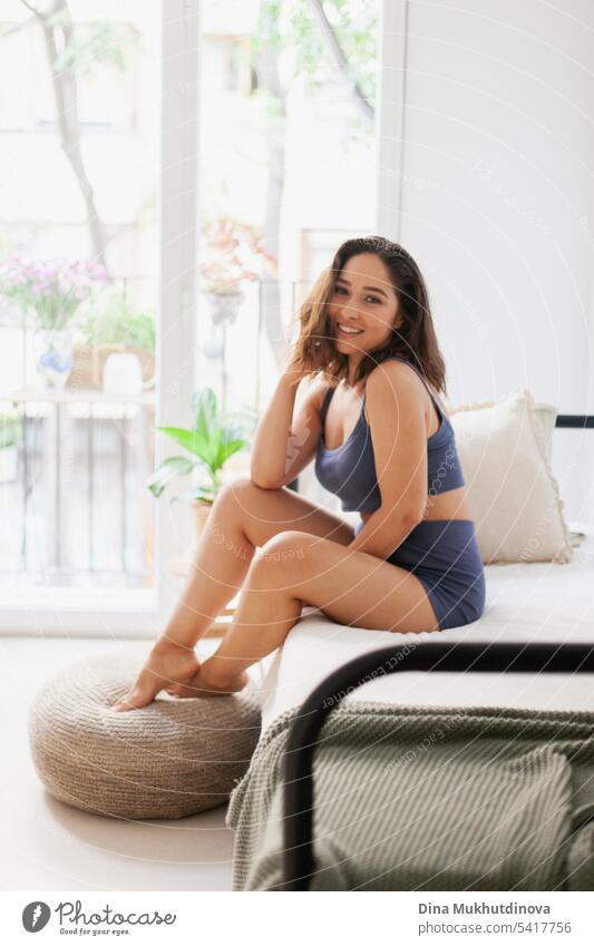 Happy young woman in minimal apartment lifestyle photo, smiling. Millennial woman at home. Interior shot with panoramic window. Girls pajama party. Good morning mood.