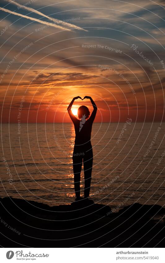 Young woman on rock shapes heart with arms at sunset by sea Sunset Sunlight sunshine Ocean Seashore Sea water seaboard Oceans coast Nature bank Summer Water