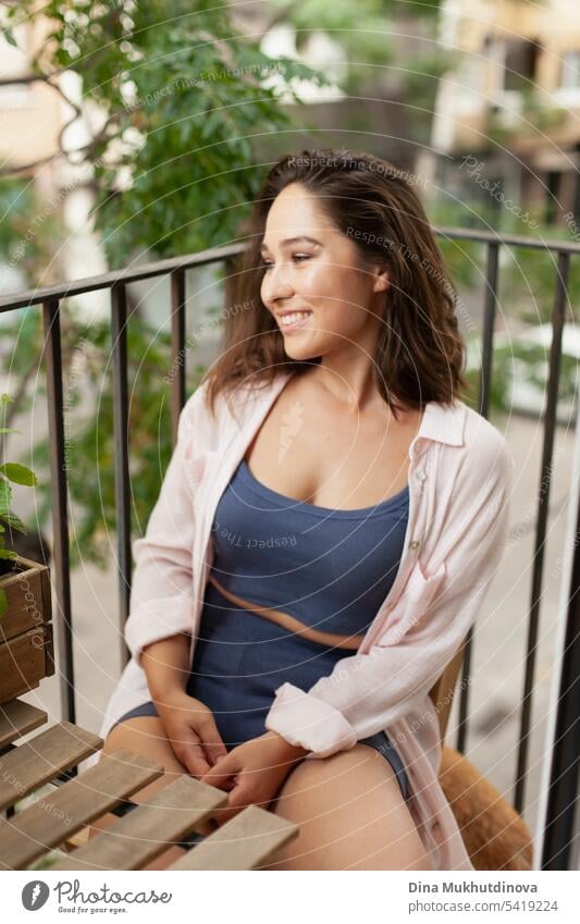 Beautiful woman  sitting on wooden chair on the balcony. Happy brunette smiling. Plants and flowers gardening hobby. Balcony plant Balcony furnishings Summer