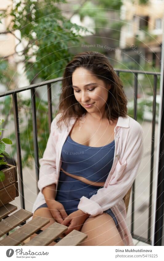 Beautiful woman  sitting on wooden chair on the balcony. Happy brunette smiling. Plants and flowers gardening hobby. Balcony plant Balcony furnishings Summer