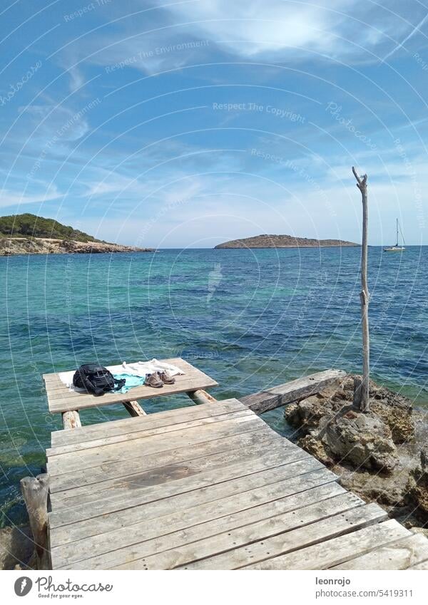 A wooden jetty overlooking the sea and waves on the Mediterranean island of Ibiza. In the background you can see a small island and a sailboat. wooden walkway