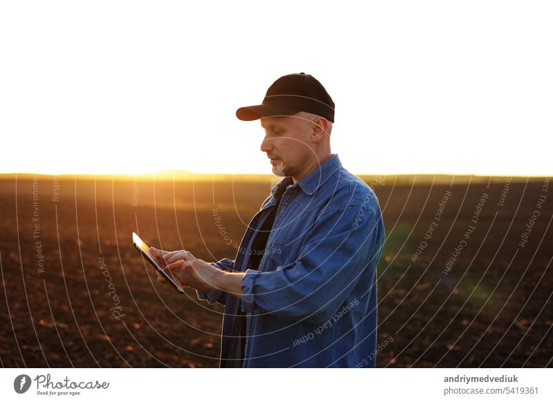 Smart farming technology and agriculture. Farmer uses digital tablet on field with plowed soil at sunset. Checking and control of soil quality, land readiness for sowing crops and planting vegetables