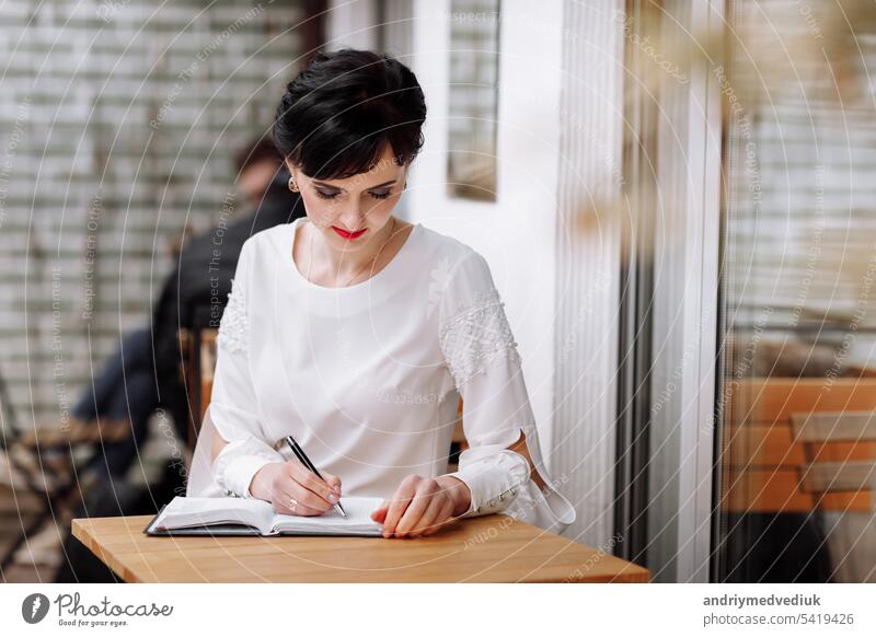 Serious focused smart businesswoman is writing notes on notepad, making agenda on personal organizer. Female in business suit sitting at table at cafe on terrace, work and event planning.