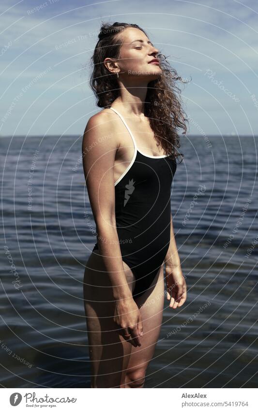 side portrait of young, beautiful, brunette, woman standing in front in bathing suit with closed eyes in shallow water of sea and full of joy pit Woman