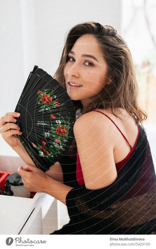 Sensual smiling woman in red and black outfit using fan. Spanish vacation. Passionate romantic female portrait in boudoir room. Summer heatwave.