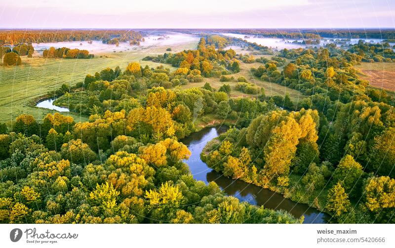 Aerial summer sunrise view. Rural landscape, river meandering in forest green trees. Morning Misty Scene. Serene atmosphere fog panorama. Agriculture fields, wood on riverbank