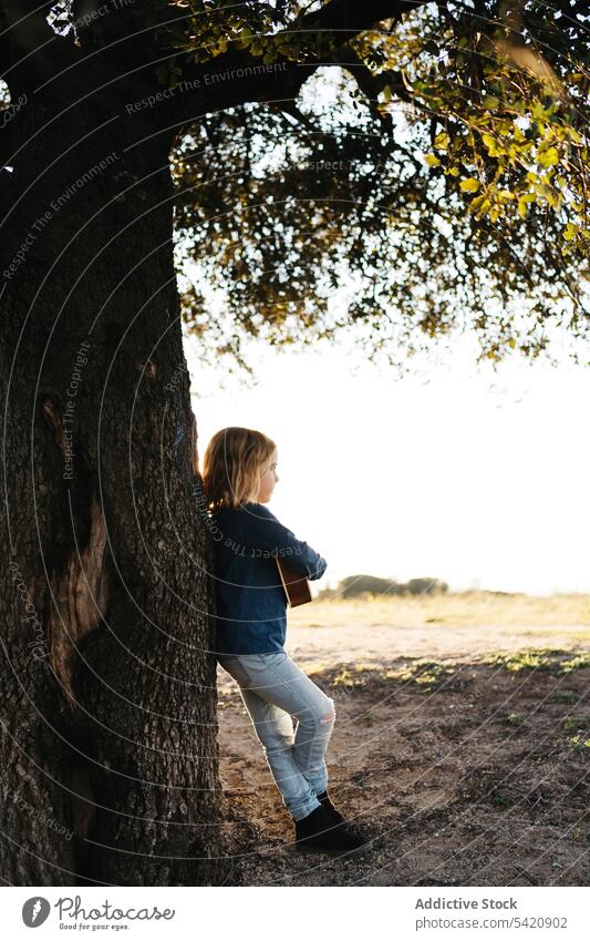 Little girl standing under tree and playing ukulele kid music guitar nature child summer serious little instrument sound song melody trunk perform practice rest