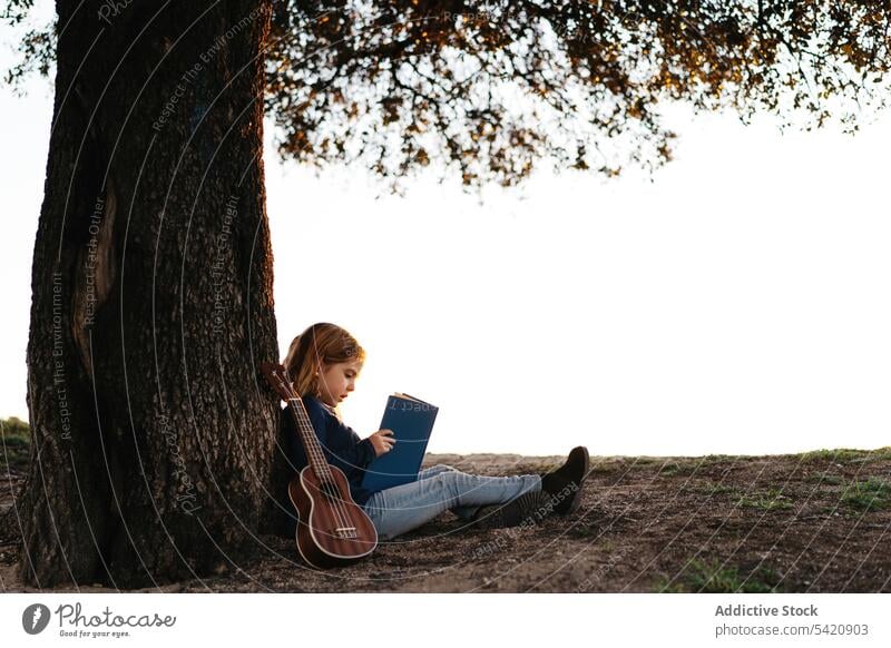 Little girl with book sitting under tree kid read nature field trunk summer casual child calm tranquil serious concentrate female learn meadow lifestyle