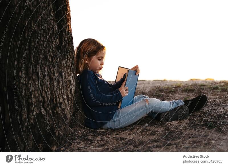 Little girl with book sitting under tree kid read nature field trunk summer casual child calm tranquil serious concentrate female learn meadow lifestyle