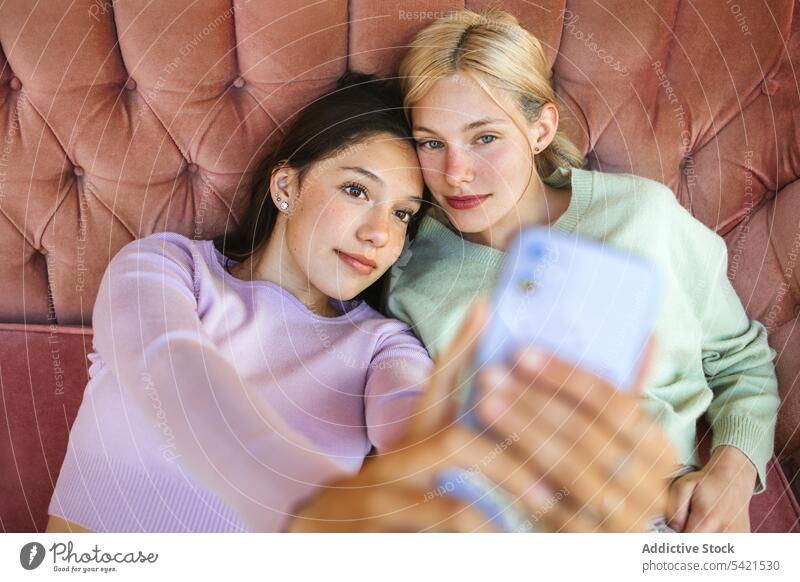 Content teenage sisters taking selfie on smartphone on couch self portrait girl mobile tender young sofa using lying device smile delicate gentle delight