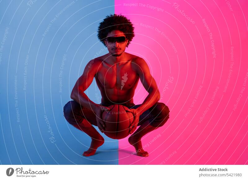 Black shirtless man sitting with basketball naked torso muscular sportsman appearance player neon squat male african american black curly hair sunglasses young