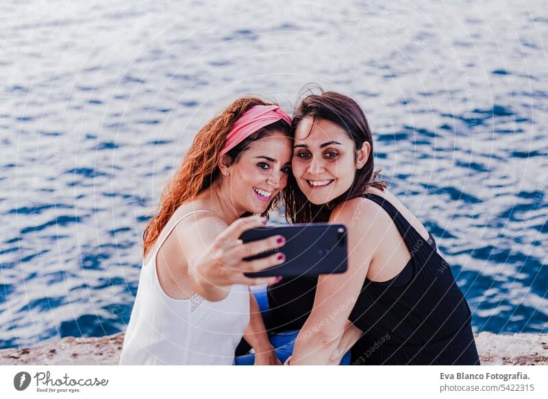 two women friends sitting by the beach using mobile phone, taking a selfie and smiling. Technology and lifestyle concept casual stylish cheerful positive