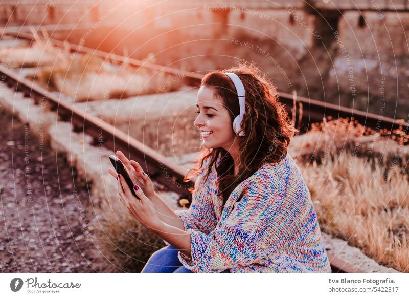 portrait outdoors of a young beautiful woman at sunset listening music on headset and smiling. Lifestyle and music concept casual fun young adult app