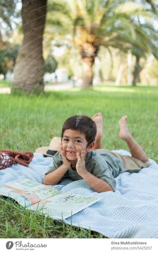 diverse kid reading a book in the park on the blue blanket on green grass, smiling and looking to the camera. Five or six years old boy in the park in summer.